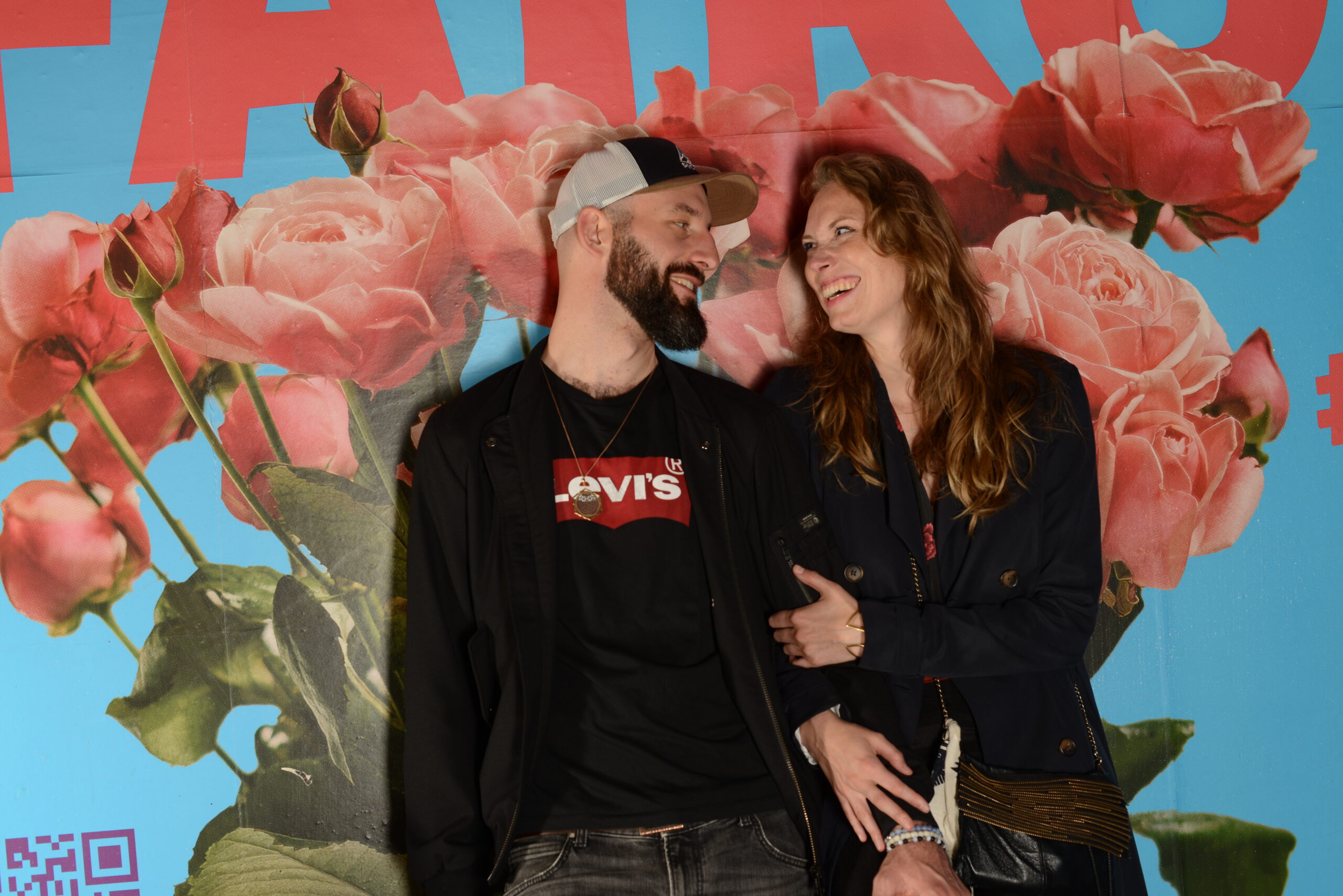 Ein glückliches Paar, das vor einem Plakat mit Rosen steht und sich anlächeln. Sie halten sich gegenseitig am Arm.